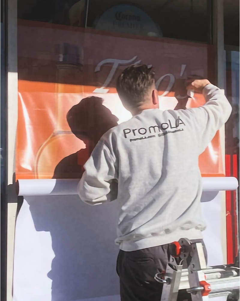 PromoLA installer in a branded sweatshirt applying a vinyl graphic to a storefront window